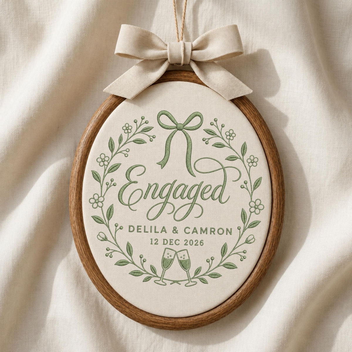 May include: An oval wooden embroidery hoop containing off-white fabric with green embroidered text reading Engaged, Delila &amp; Camron, and 12 Dec 2026. The design features a green floral wreath, a bow, and two champagne flutes, topped with a beige ribbon bow.