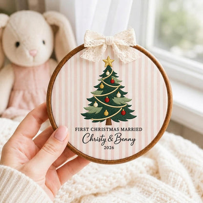 May include: A circular wooden embroidery hoop frame containing fabric with a light pink and white vertical stripe pattern. The fabric features an embroidered green Christmas tree with red and gold ornaments, topped with a gold star. Text reads: FIRST CHRISTMAS MARRIED, Christy &amp; Benny, 2026. A cream lace bow is attached to the top.