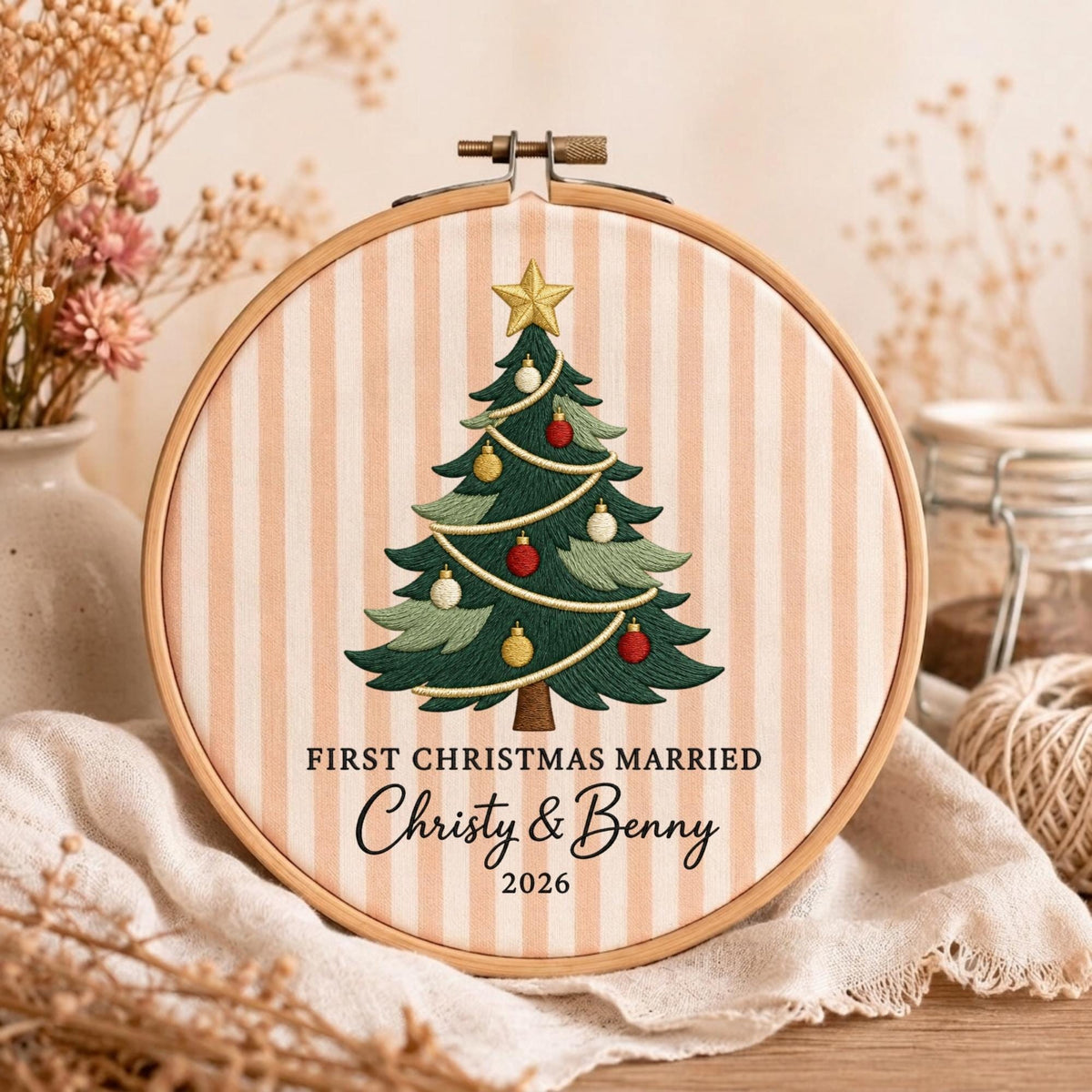 May include: A round wooden embroidery hoop containing fabric with vertical peach and white stripes. The fabric features an embroidered green Christmas tree with red, gold, and white ornaments, topped with a gold star. Text reads: FIRST CHRISTMAS MARRIED, Christy &amp; Benny, 2026.