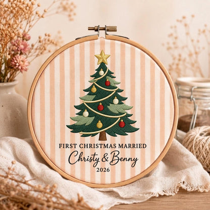 May include: A round wooden embroidery hoop containing fabric with vertical peach and white stripes. The fabric features an embroidered green Christmas tree with red, gold, and white ornaments, topped with a gold star. Text reads: FIRST CHRISTMAS MARRIED, Christy &amp; Benny, 2026.