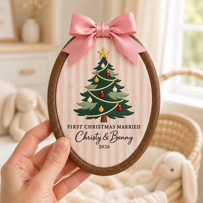 May include: An oval wooden embroidery hoop frame containing fabric with a pink and white striped pattern. An embroidered green Christmas tree with gold, red, and white ornaments is centered. Text reads: FIRST CHRISTMAS MARRIED Christy &amp; Benny 2026.