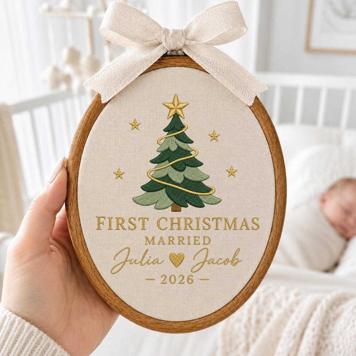 May include: An oval wooden embroidery hoop containing cream-colored fabric with an embroidered green Christmas tree, gold stars, and the text "FIRST CHRISTMAS MARRIED Julia Jacob 2026" with a gold heart. A beige ribbon bow is tied at the top.