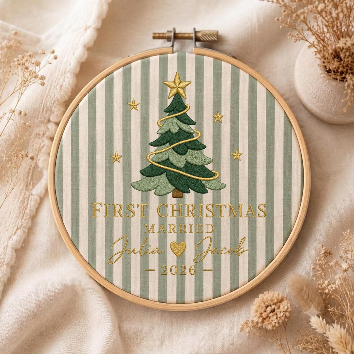May include: A circular wooden embroidery hoop framing fabric with sage green and white vertical stripes. Centered is an embroidered green Christmas tree with a gold star and garland, surrounded by gold stars and the text FIRST CHRISTMAS MARRIED Julia and Jacob 2026.