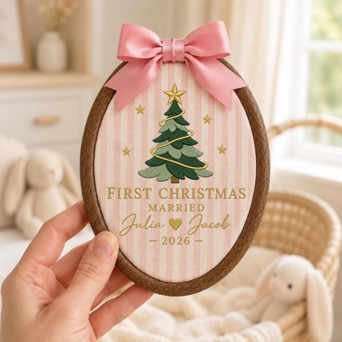May include: An oval wooden embroidery hoop frame containing fabric with vertical pink and white stripes. The fabric features an embroidered green Christmas tree with a gold star, gold text reading FIRST CHRISTMAS MARRIED Julia and Jacob 2026, and a pink satin bow.