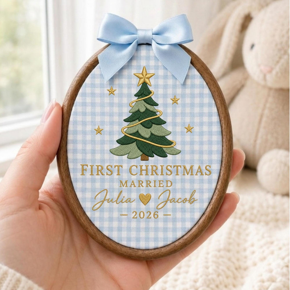 May include: An oval wooden embroidery hoop containing fabric with a light blue and white gingham pattern. The fabric features an embroidered green Christmas tree with a gold star, gold stars, and the text FIRST CHRISTMAS MARRIED Julia Jacob 2026.