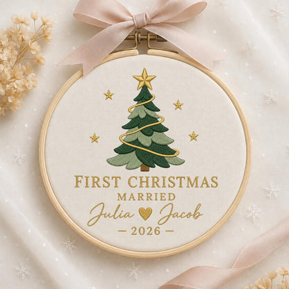 May include: A round wooden embroidery hoop containing white fabric with a stitched green Christmas tree, gold stars, and the text FIRST CHRISTMAS MARRIED Julia and Jacob 2026. A light pink ribbon is tied to the top of the wooden frame.