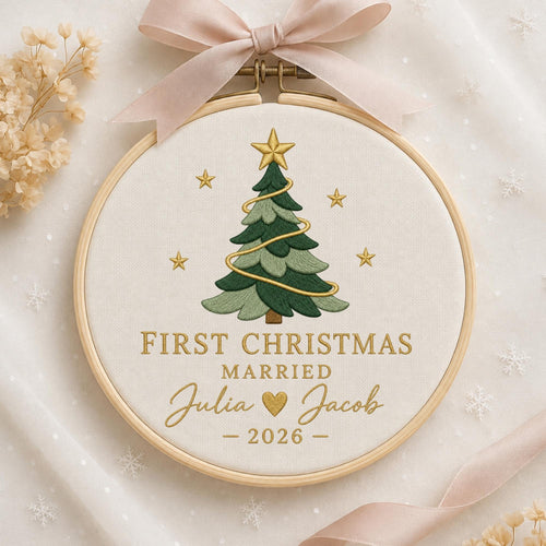 May include: A round wooden embroidery hoop containing white fabric with a stitched green Christmas tree, gold stars, and the text FIRST CHRISTMAS MARRIED Julia and Jacob 2026. A light pink ribbon is tied to the top of the wooden frame.