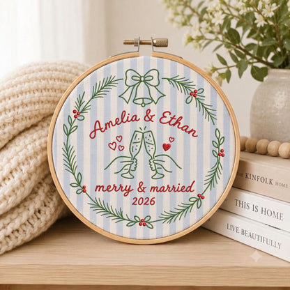 May include: A circular embroidery hoop featuring light blue and white striped fabric. The design includes a green pine wreath with red berries, a bell, and two hands clinking champagne glasses. Text reads: Amelia &amp; Ethan, merry &amp; married, 2026.
