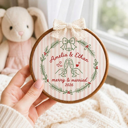 May include: A circular wooden embroidery hoop containing fabric with pink and white vertical stripes. The design features a green pine wreath, a bell, and two hands clinking champagne glasses with the text Amelia &amp; Ethan merry &amp; married 2026.