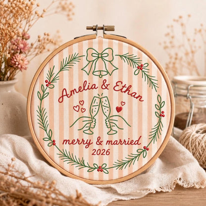 May include: A round wooden embroidery hoop containing fabric with vertical beige stripes. The embroidery features a green wreath with red berries, a bow, two hands clinking champagne glasses, and the text Amelia &amp; Ethan merry &amp; married 2026.