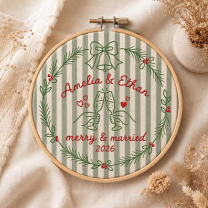 May include: A circular embroidery hoop containing fabric with green and white vertical stripes. The design features a green wreath with red berries, a bow, and two champagne flutes clinking. Text reads: Amelia &amp; Ethan, merry &amp; married, 2026.