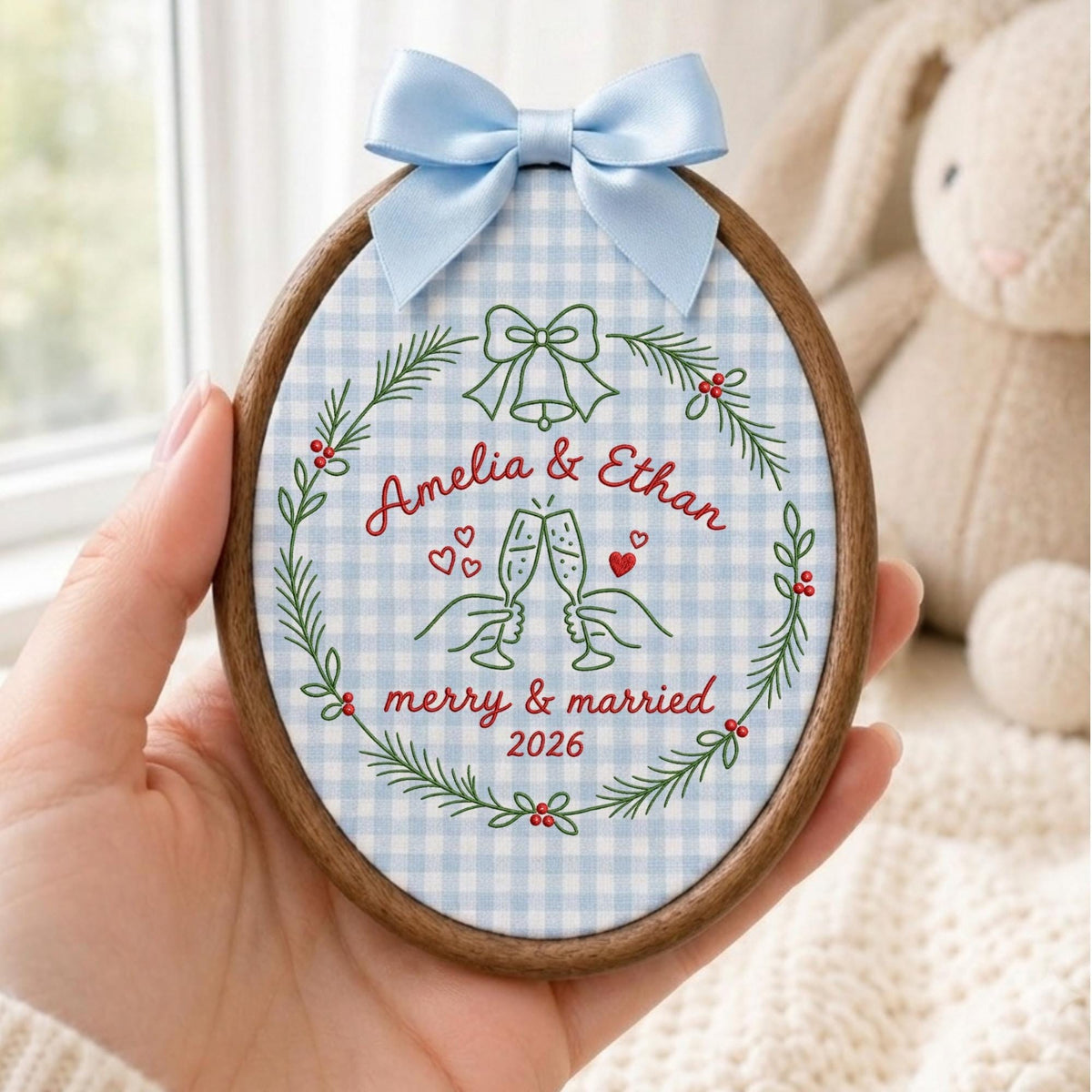 May include: An oval wooden embroidery hoop containing light blue and white gingham fabric. The fabric features embroidered green pine branches, red berries, a bell, two hands clinking champagne glasses, and the text Amelia &amp; Ethan merry &amp; married 2026. A light blue satin ribbon bow is attached to the top.