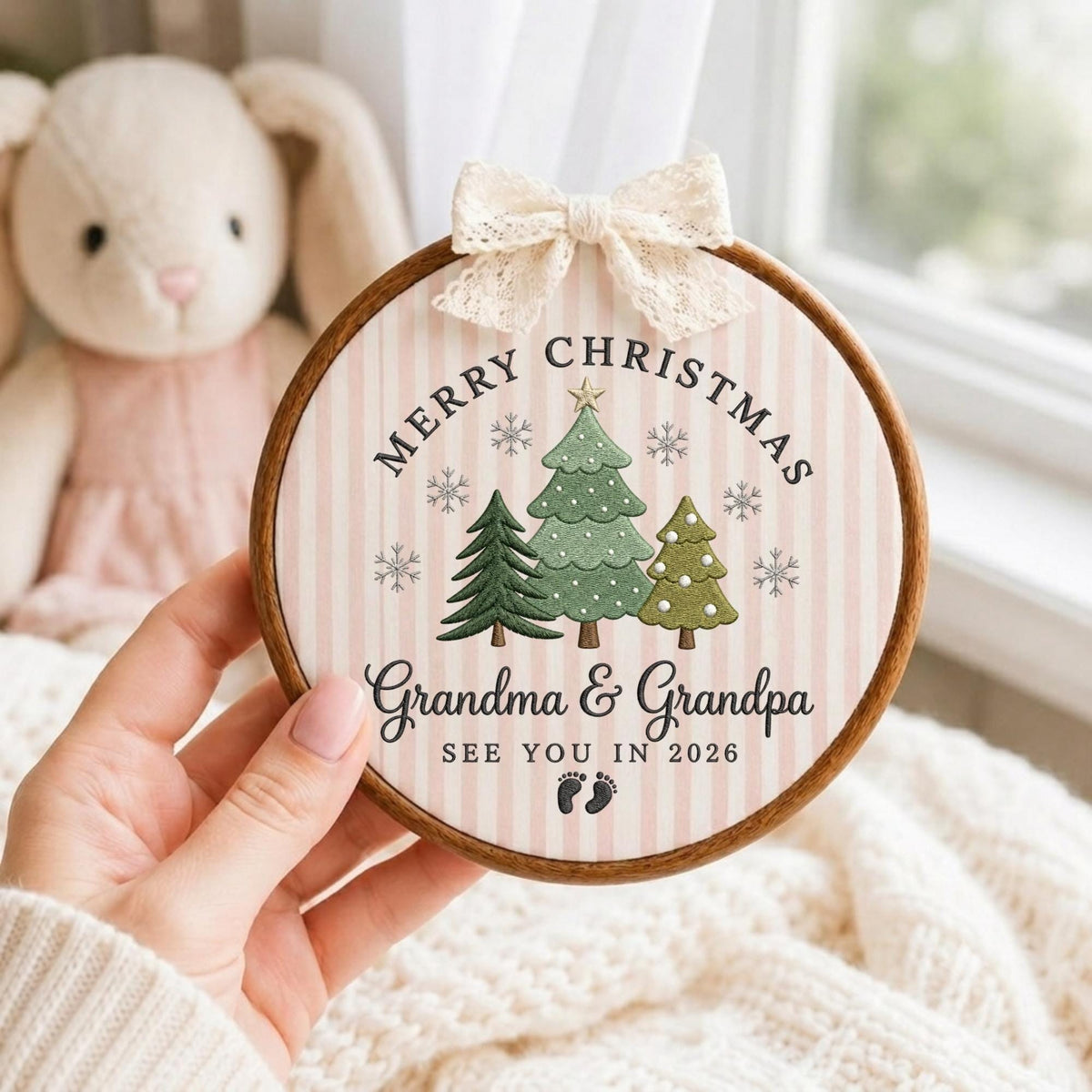 May include: A round wooden embroidery hoop ornament featuring a light pink and white striped fabric background. The design includes three green Christmas trees, snowflakes, and the text Merry Christmas Grandma &amp; Grandpa SEE YOU IN 2026 with baby footprints. A cream lace bow is attached to the top.