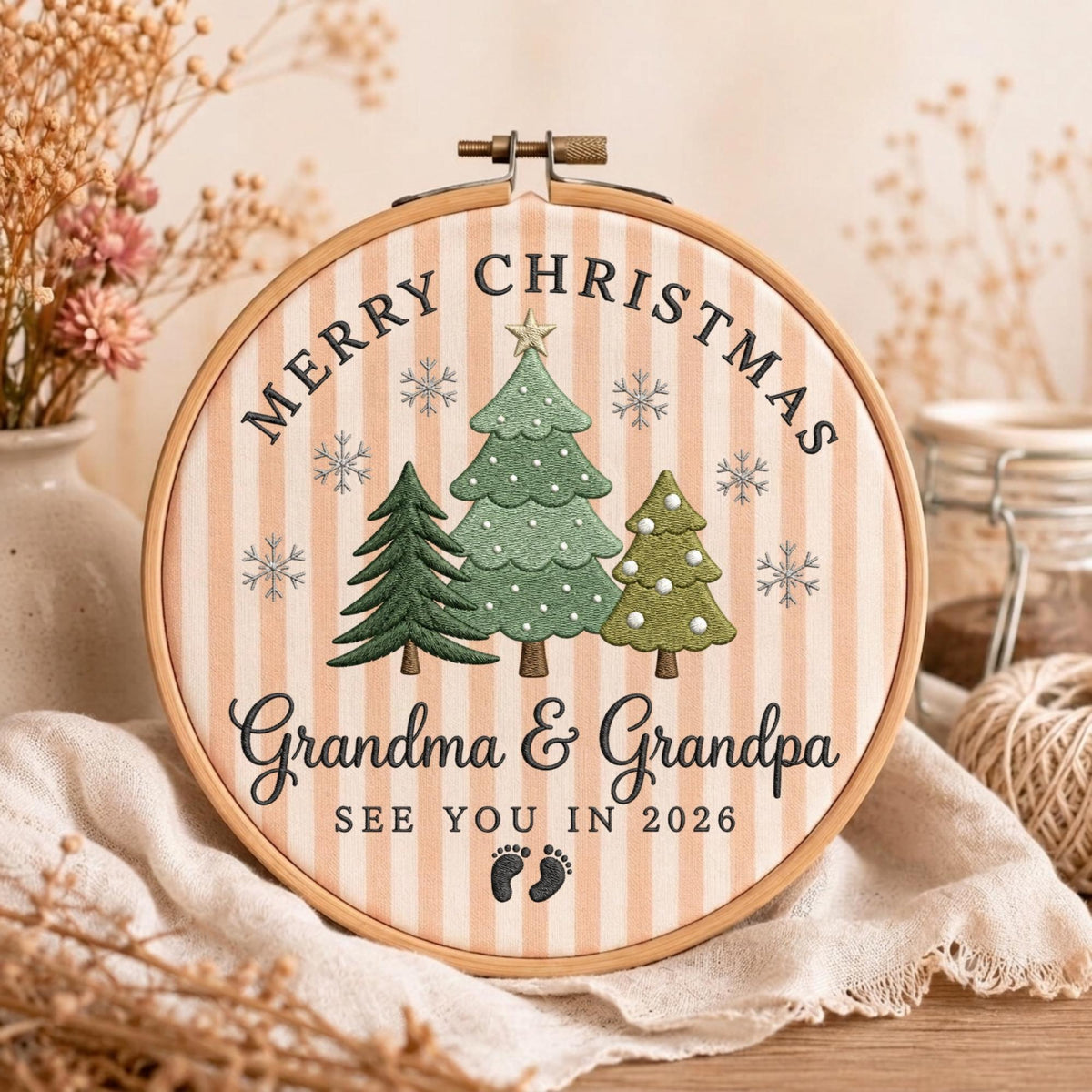 May include: A round wooden embroidery hoop containing fabric with vertical beige stripes. The design features three embroidered green Christmas trees, snowflakes, and the text Merry Christmas, Grandma &amp; Grandpa, SEE YOU IN 2026, and two small footprints.