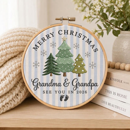 May include: A circular embroidery hoop featuring a light blue and white striped fabric background. The design includes three embroidered green Christmas trees, snowflakes, and the text "MERRY CHRISTMAS Grandma &amp; Grandpa SEE YOU IN 2026" with two small black baby footprints.