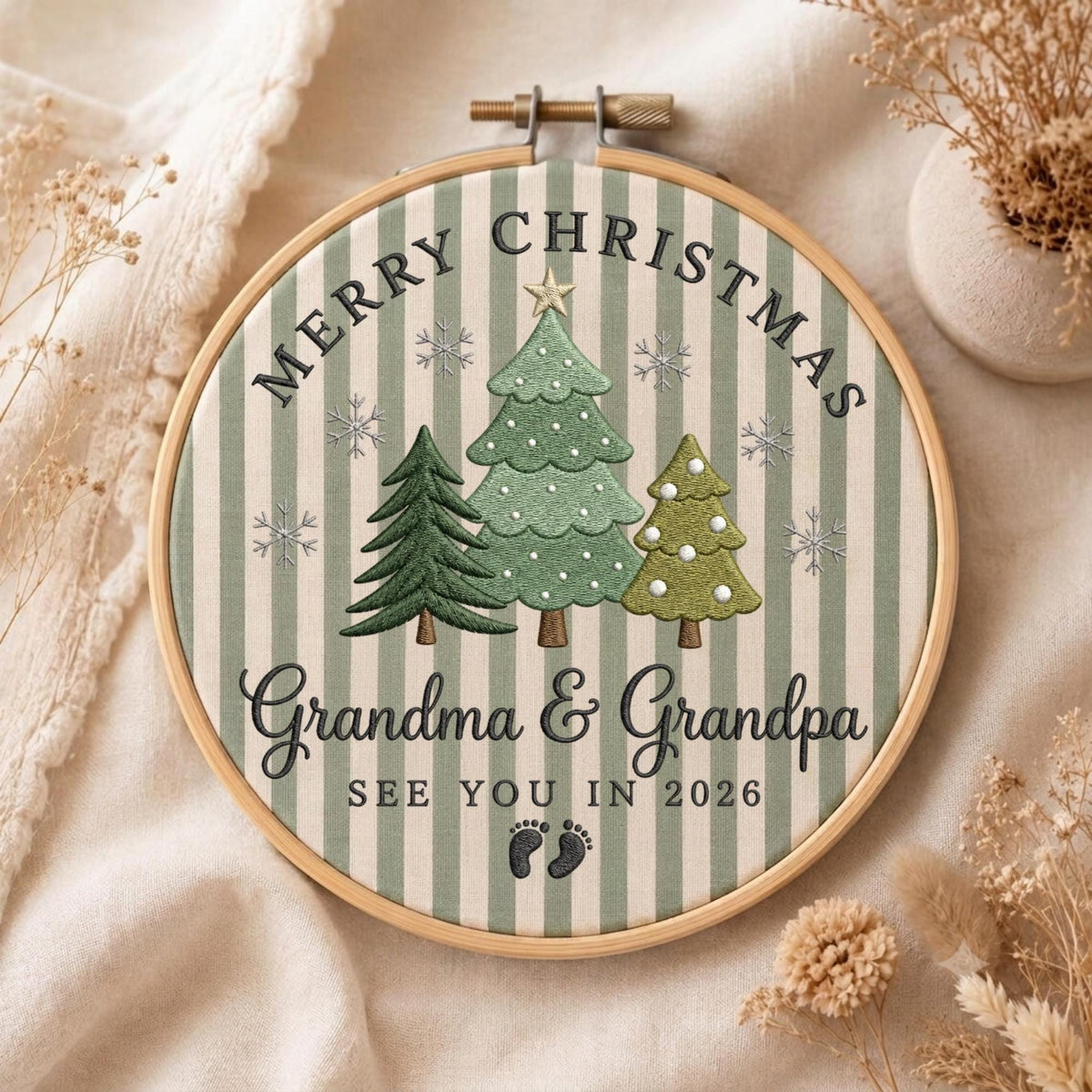 May include: A circular embroidery hoop featuring a light green and white striped fabric background. The design includes three embroidered Christmas trees, snowflakes, and the text Merry Christmas, Grandma &amp; Grandpa, SEE YOU IN 2026, and baby footprints.