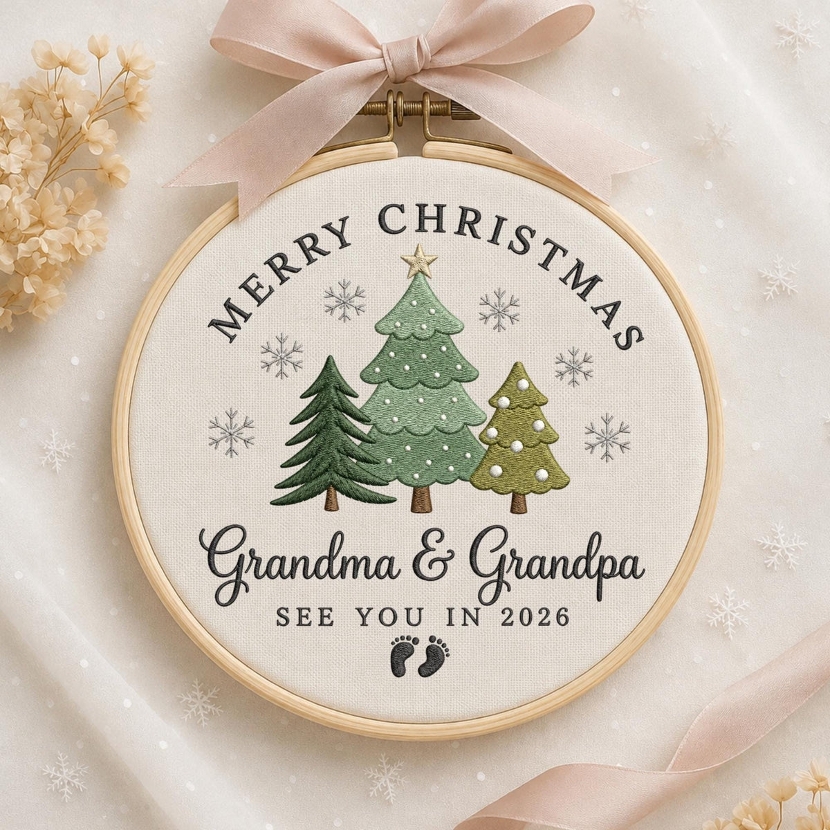 May include: A circular wooden embroidery hoop containing white fabric with stitched green Christmas trees, snowflakes, and the text Merry Christmas Grandma &amp; Grandpa SEE YOU IN 2026 with two small black footprints, topped with a light pink ribbon.