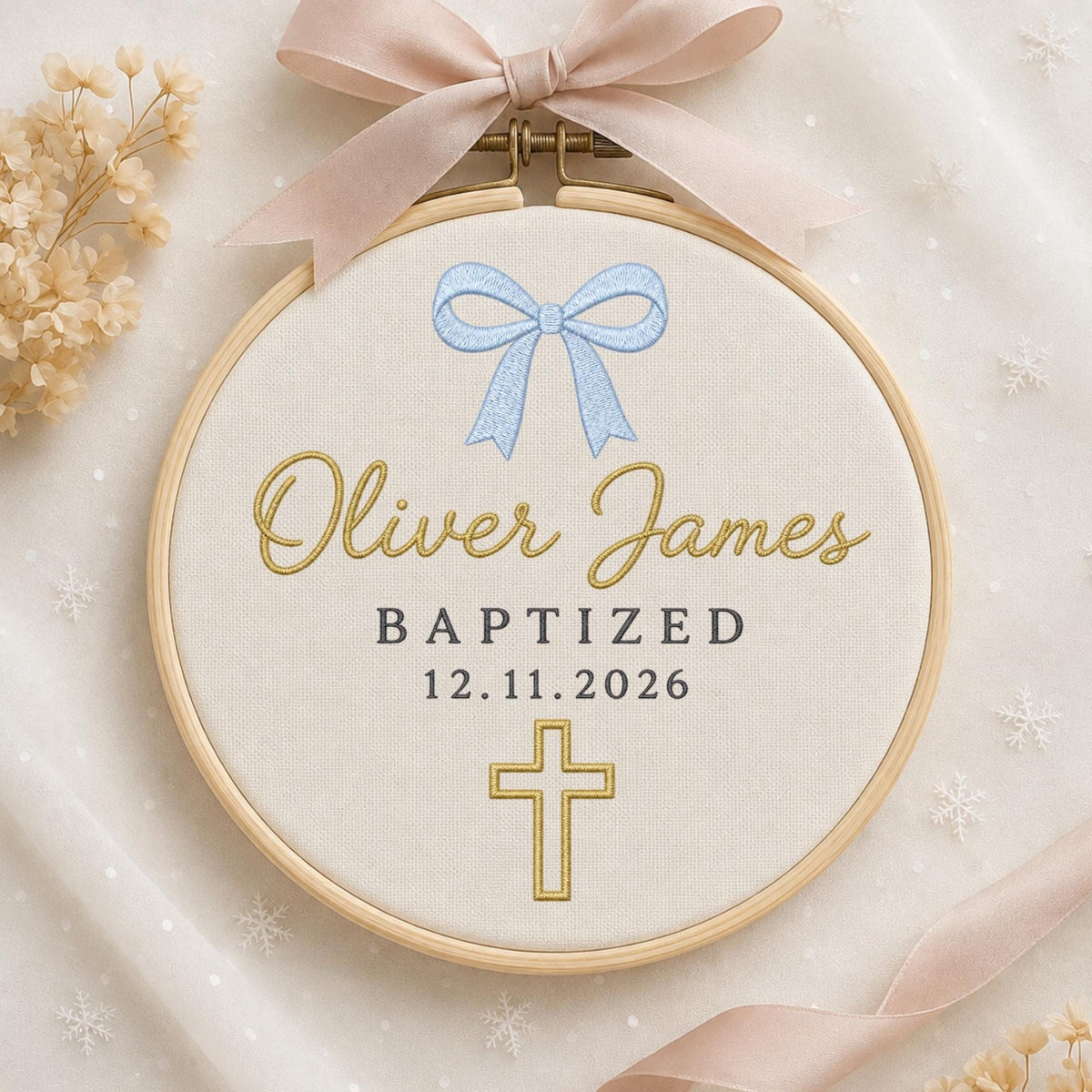 May include: A circular wooden embroidery hoop containing cream fabric with a light blue embroidered bow, the gold text Oliver James, the word BAPTIZED, the date 12.11.2026, and a gold cross. A large pale pink ribbon bow is attached to the top.