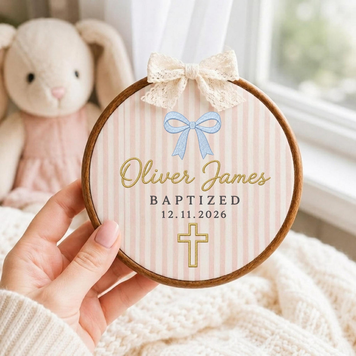May include: A circular wooden embroidery hoop frame containing fabric with a light pink and white vertical striped pattern. The fabric features a blue bow graphic, the text Oliver James, BAPTIZED, 12.11.2026, a gold cross, and a cream lace bow.