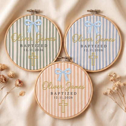 May include: Three circular wooden embroidery hoops featuring striped fabric in sage green, light blue, and peach. Each hoop displays an embroidered light blue bow, the name Oliver James, the text BAPTIZED 12.11.2026, and a gold-stitched cross.