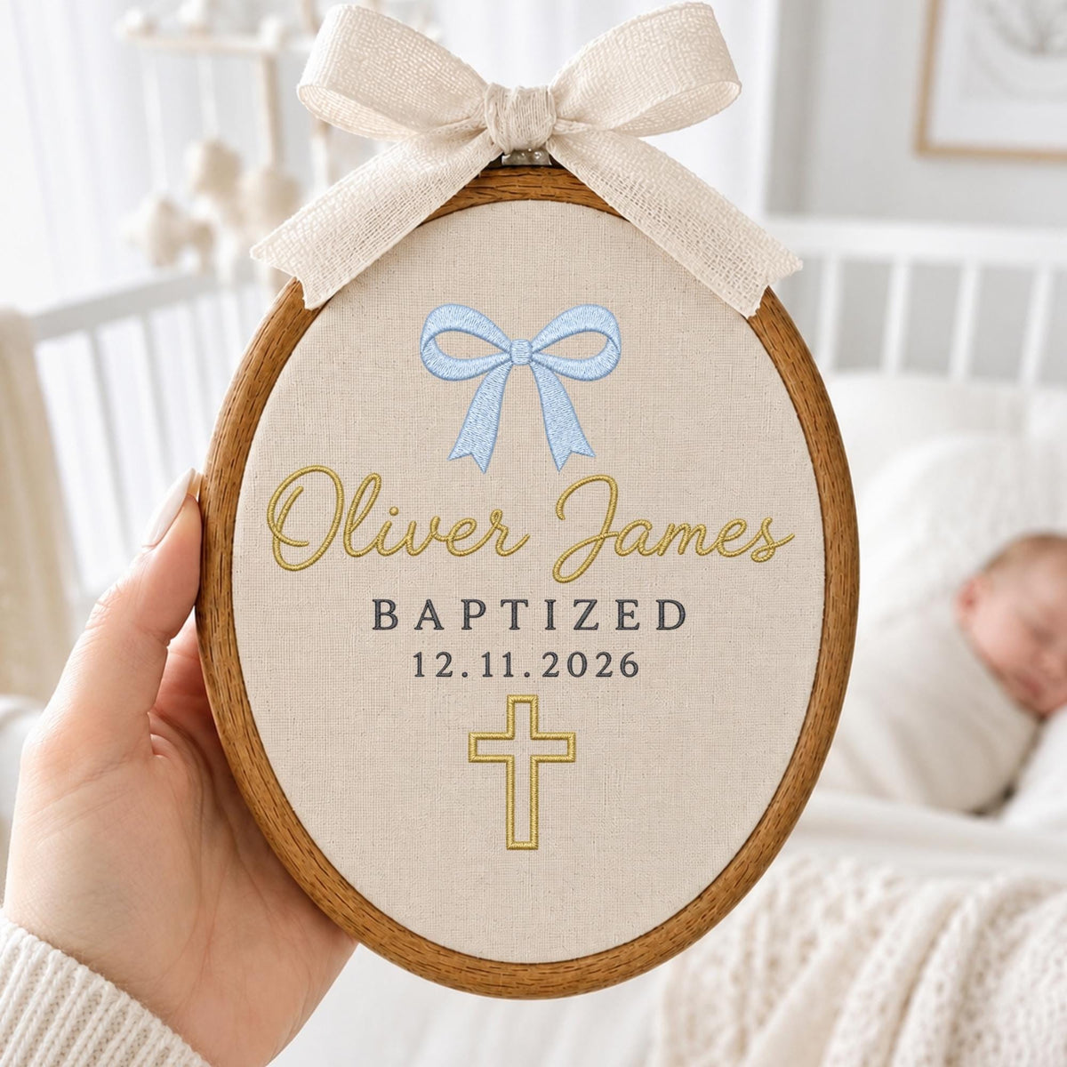 May include: An oval wooden embroidery hoop containing beige fabric with a light blue embroidered bow, the gold text "Oliver James", the dark text "BAPTIZED 12.11.2026", and a gold embroidered cross. A beige fabric bow is tied to the top of the hoop.
