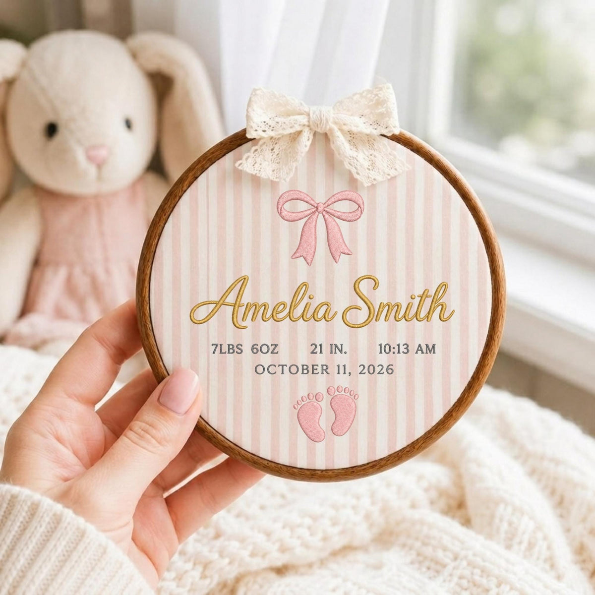 May include: A circular wooden embroidery hoop featuring a light pink and white striped fabric background. The design includes a cream lace bow at the top, a pink bow graphic, the name Amelia Smith in gold, and birth details: 7LBS 6OZ, 21 IN., 10:13 AM, OCTOBER 11, 2026, with two small pink baby footprints.