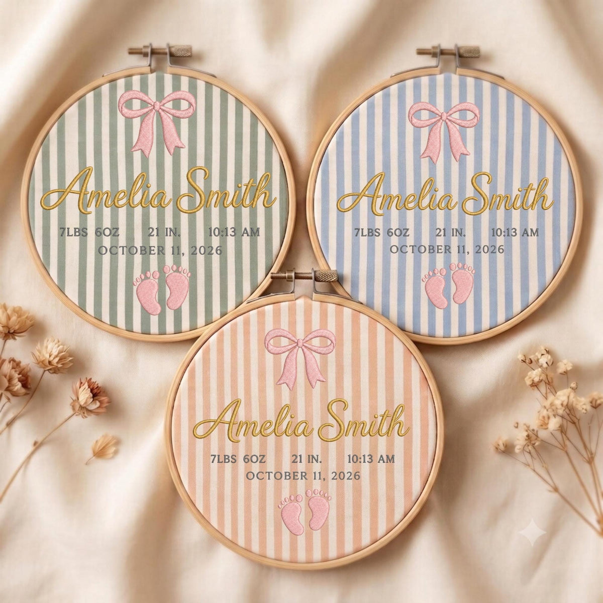 May include: Three circular embroidery hoops featuring striped fabric in sage green, light blue, and peach. Each displays the name Amelia Smith in gold script, a pink bow, baby footprint icons, and birth details: 7lbs 6oz, 21 in, 10:13 am, October 11, 2026.