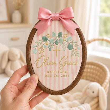 May include: An oval wooden embroidery hoop containing fabric with pink and white vertical stripes. The fabric features an embroidered green eucalyptus wreath, a gold cross, and the text Olivia Grace BAPTIZED DEC 25, 2026. A pink satin bow is attached to the top.