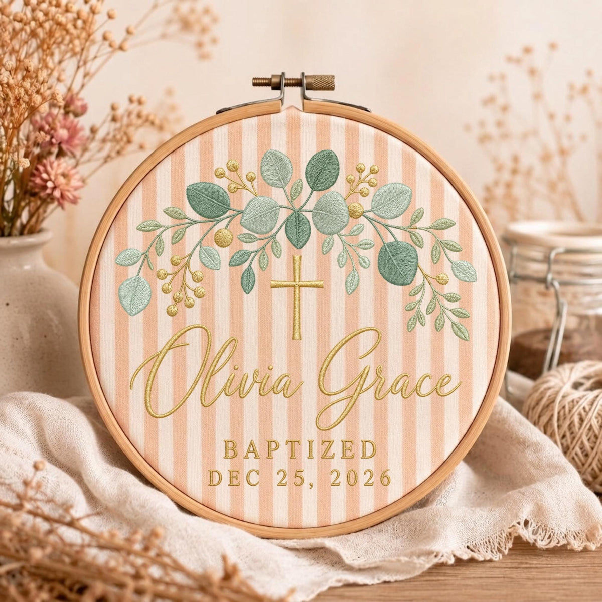 May include: A circular wooden embroidery hoop framing a light peach and white striped fabric. The fabric features an embroidered green eucalyptus wreath, a gold cross, and the text Olivia Grace, BAPTIZED DEC 25, 2026 in gold thread.