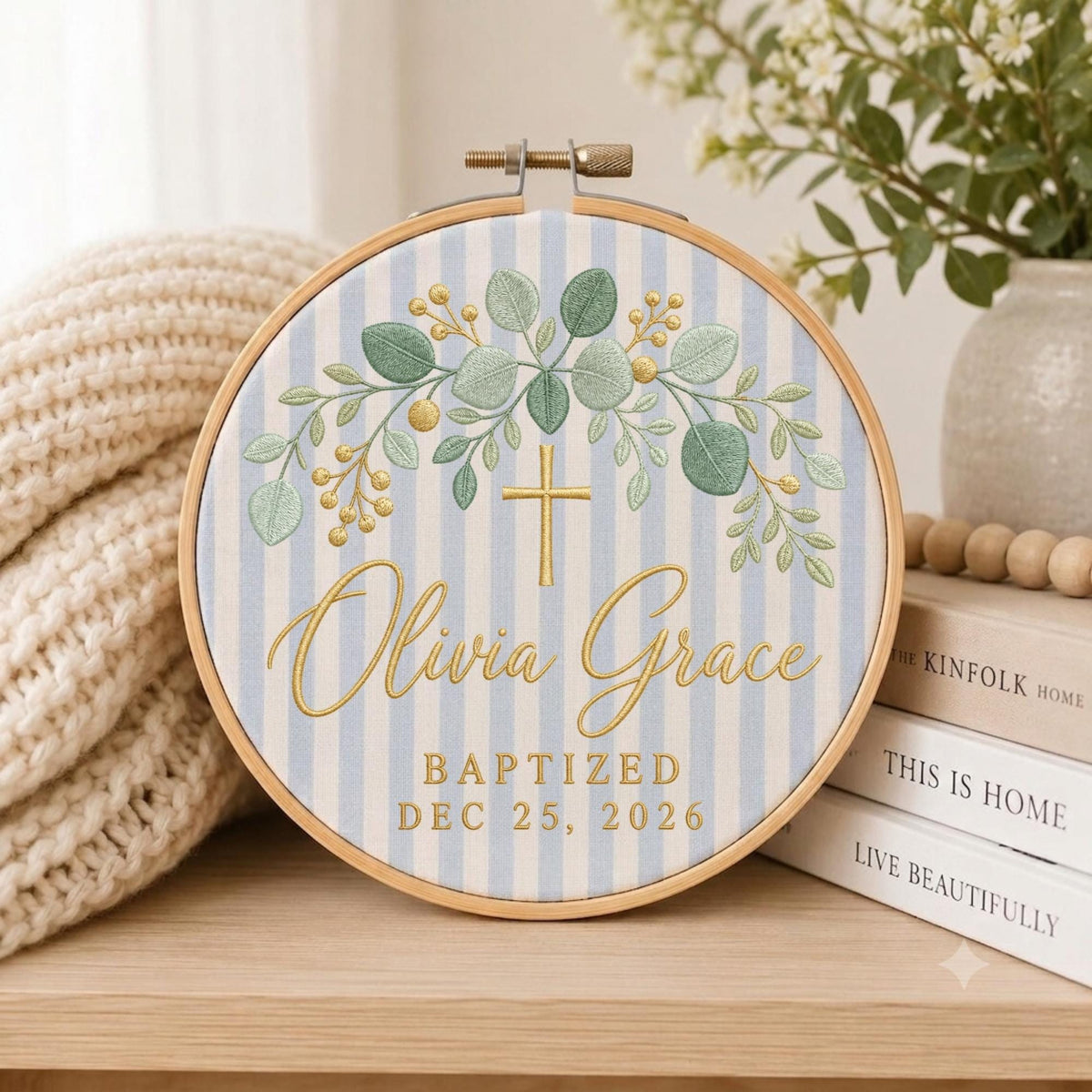 May include: A circular wooden embroidery hoop containing fabric with light blue and white vertical stripes. The design features embroidered green eucalyptus leaves, gold berries, a gold cross, and the text Olivia Grace BAPTIZED DEC 25, 2026.