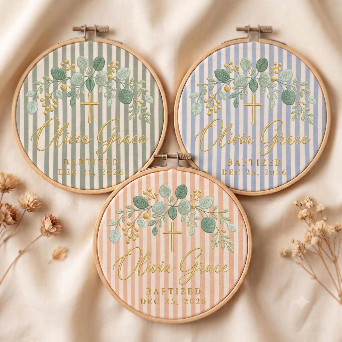 May include: Three circular wooden embroidery hoops featuring fabric with vertical stripes in sage green, light blue, and peach. Each hoop displays embroidered green eucalyptus leaves, a gold cross, and the text Olivia Grace Baptized Dec 25, 2026.