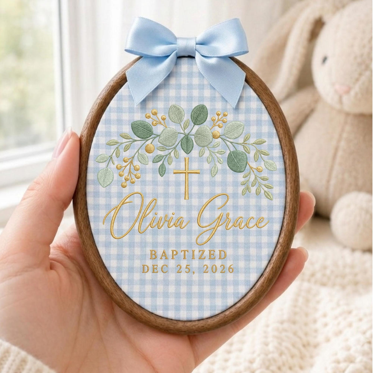 May include: An oval wooden embroidery hoop frame containing fabric with a light blue and white gingham pattern. It features a gold cross, green eucalyptus leaves, and the text Olivia Grace, BAPTIZED, and DEC 25, 2026. A light blue satin bow is attached.