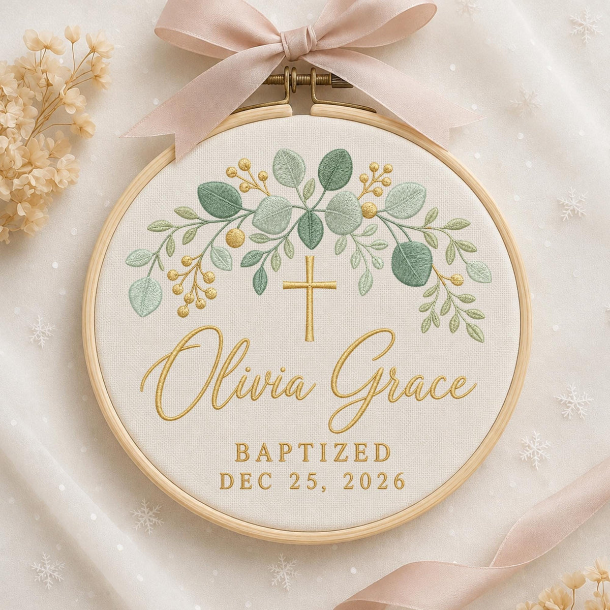 May include: A circular wooden embroidery hoop containing white fabric with an embroidered design of green eucalyptus leaves and gold berries. A gold cross is centered above the name Olivia Grace and the text BAPTIZED DEC 25, 2026. A pink ribbon is tied at the top.