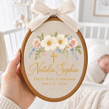 May include: An oval wooden embroidery hoop containing beige fabric with embroidered white and pink flowers, green leaves, a gold cross, and the text "Natalia Sophia", "First Holy Communion", and "Sept 11, 2026". A beige ribbon bow is tied at the top.