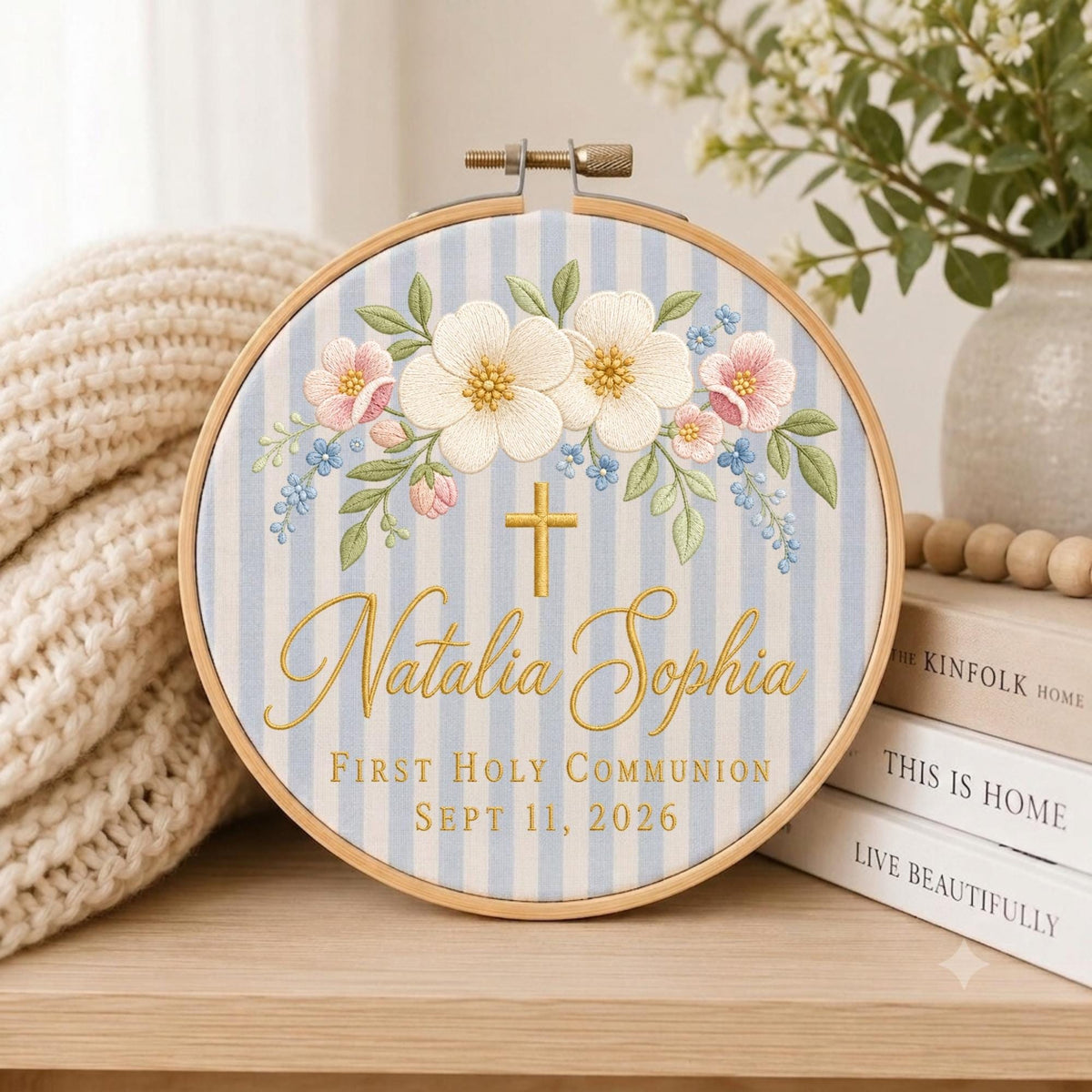 May include: A circular embroidery hoop featuring a light blue and white striped fabric background. The design includes embroidered white and pink flowers with green leaves, a gold cross, and the text Natalia Sophia First Holy Communion Sept 11, 2026.