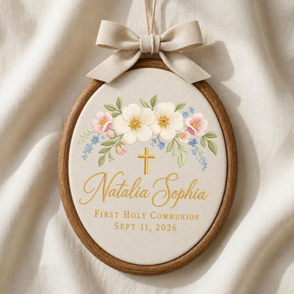 May include: An oval wooden embroidery hoop containing cream fabric with a floral design, a gold cross, and the text Natalia Sophia First Holy Communion Sept 11, 2026. A beige fabric bow is attached to the top of the hoop for hanging purposes.