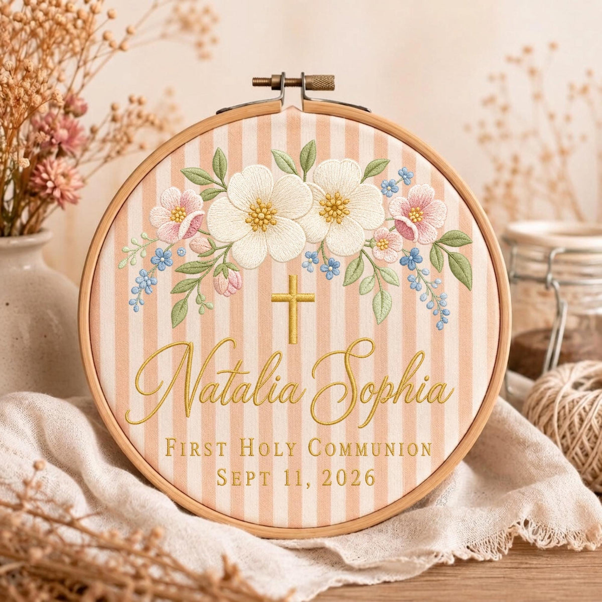 May include: A round wooden embroidery hoop containing fabric with vertical light pink and white stripes. The fabric features embroidered white and pink flowers, green leaves, a gold cross, and the text Natalia Sophia First Holy Communion Sept 11, 2026.
