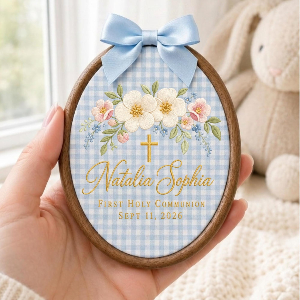 May include: An oval wooden embroidery hoop frame containing fabric with a light blue gingham pattern. The fabric features embroidered white and pink flowers, a gold cross, and the text Natalia Sophia, FIRST HOLY COMMUNION, SEPT 11, 2026. A light blue satin bow is attached to the top.