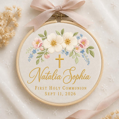 May include: A circular wooden embroidery hoop containing off-white fabric with floral embroidery in white, pink, and blue. A gold cross is stitched above the name Natalia Sophia, followed by the text FIRST HOLY COMMUNION SEPT 11, 2026. A light pink ribbon is tied at the top.