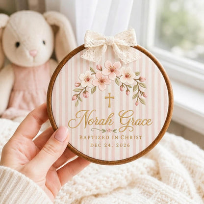 May include: A circular wooden embroidery hoop frame containing fabric with a pink and white vertical stripe pattern. The design features a floral arch, a gold cross, and the text Norah Grace, BAPTIZED IN CHRIST, DEC 24, 2026. A cream lace bow is attached to the top.