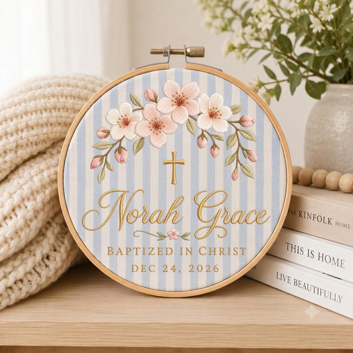 May include: A circular embroidery hoop featuring light blue and white vertical stripes. The fabric displays an embroidered floral arch of pink cherry blossoms above a gold cross. The text reads Norah Grace, BAPTIZED IN CHRIST, DEC 24, 2026.