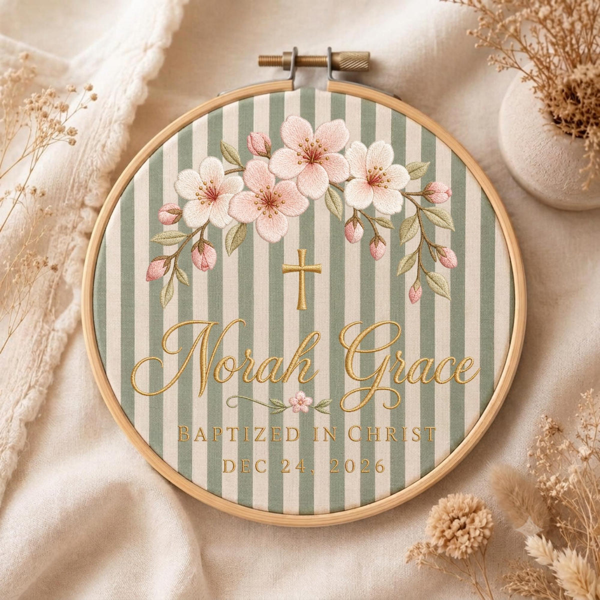 May include: A circular wooden embroidery hoop containing fabric with sage green and white vertical stripes. The design features embroidered pink cherry blossoms, a gold cross, and the text Norah Grace, BAPTIZED IN CHRIST, DEC 24, 2026.