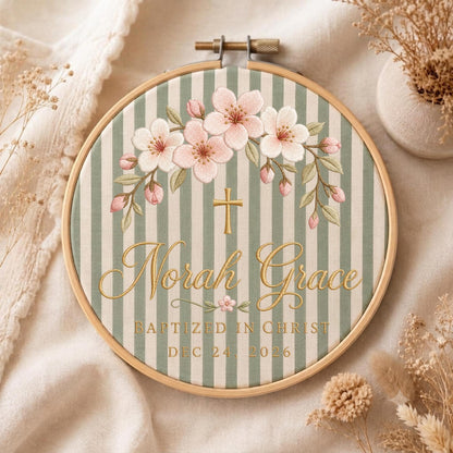 May include: A circular wooden embroidery hoop containing fabric with sage green and white vertical stripes. The design features embroidered pink cherry blossoms, a gold cross, and the text Norah Grace, BAPTIZED IN CHRIST, DEC 24, 2026.