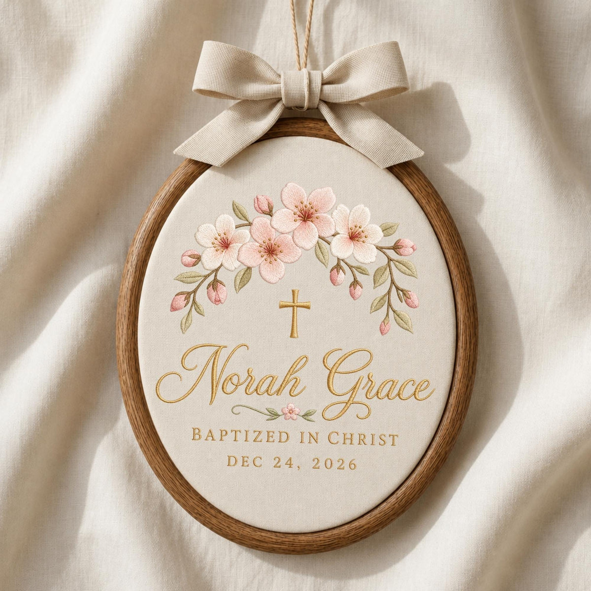 May include: An oval wooden embroidery hoop containing cream fabric with a beige ribbon bow at the top. The design features a floral arch of pink cherry blossoms above a gold cross, the name Norah Grace, and the text BAPTIZED IN CHRIST DEC 24, 2026.
