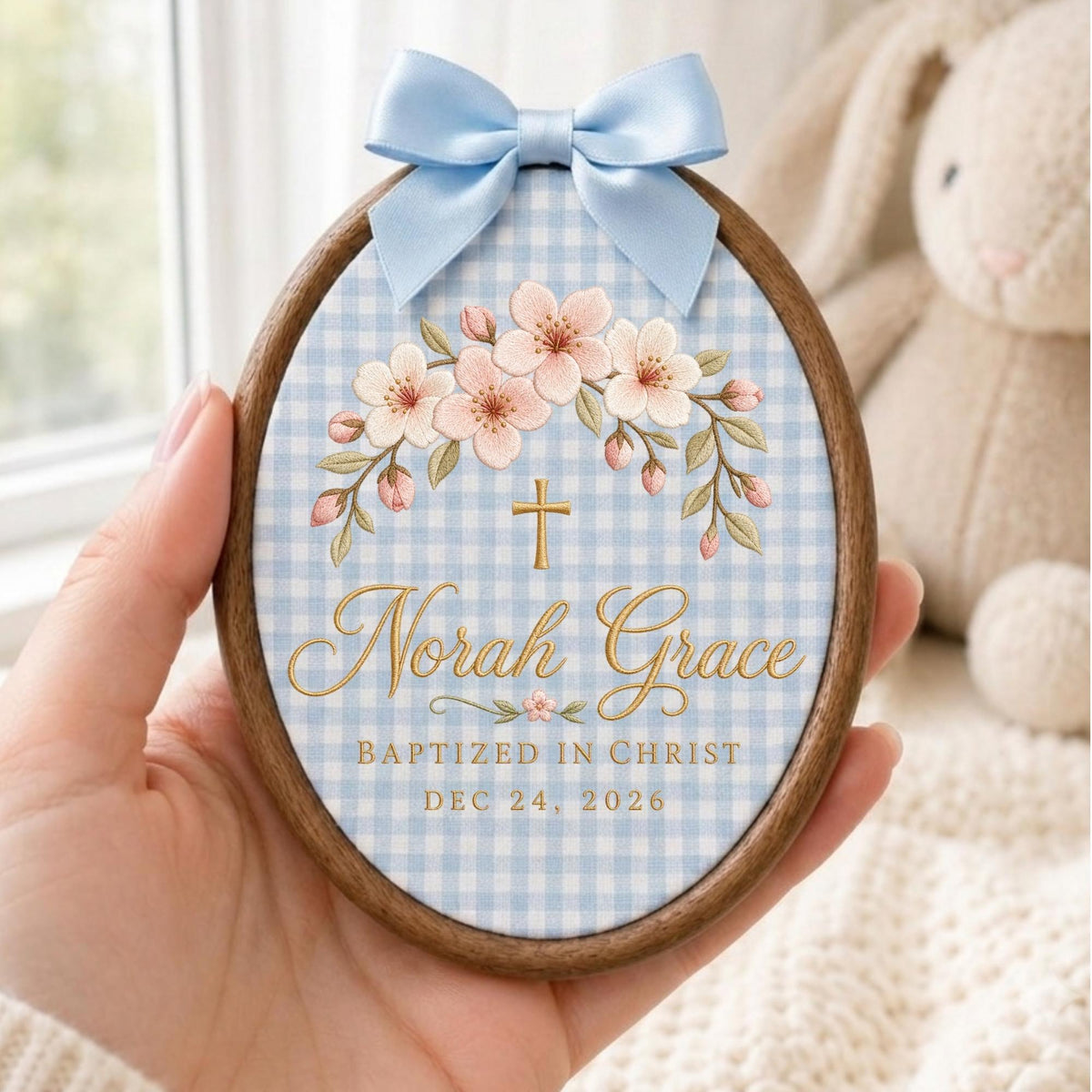 May include: An oval wooden embroidery hoop frame containing fabric with a light blue gingham pattern. It features a light blue satin bow at the top, a floral arch of pink blossoms, a gold cross, and the text Norah Grace, BAPTIZED IN CHRIST, DEC 24, 2026.