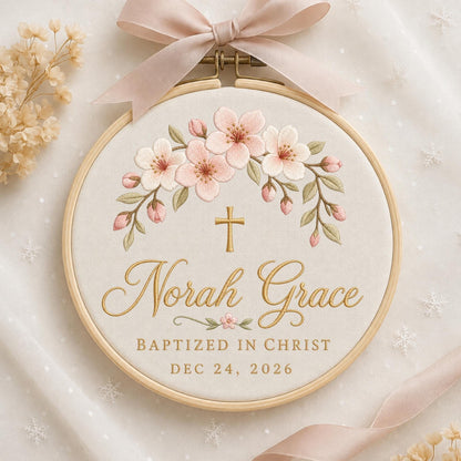 May include: A circular wooden embroidery hoop containing cream fabric with an embroidered arch of pink cherry blossoms and green leaves. A gold cross sits above the gold cursive text Norah Grace, BAPTIZED IN CHRIST, and DEC 24, 2026.