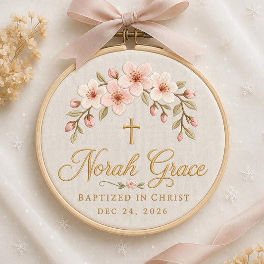 May include: A circular wooden embroidery hoop containing cream fabric with an embroidered arch of pink cherry blossoms and green leaves. A gold cross sits above the gold cursive text Norah Grace, BAPTIZED IN CHRIST, and DEC 24, 2026.