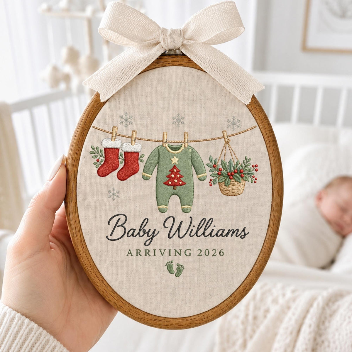 May include: An oval wooden embroidery hoop containing a beige fabric panel with a festive design. The embroidery features two red stockings, a green baby onesie with a Christmas tree, and a basket of holly hanging on a line with snowflakes. Text reads Baby Williams ARRIVING 2026 with small footprints. A beige ribbon bow is attached to the top.
