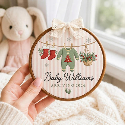 May include: A round wooden embroidery hoop featuring a light pink and white striped fabric background. The design includes two red stockings, a green baby onesie with a red Christmas tree, and a basket of holly hanging on a line. Text reads Baby Williams ARRIVING 2026 with two small green footprints and a cream lace bow.