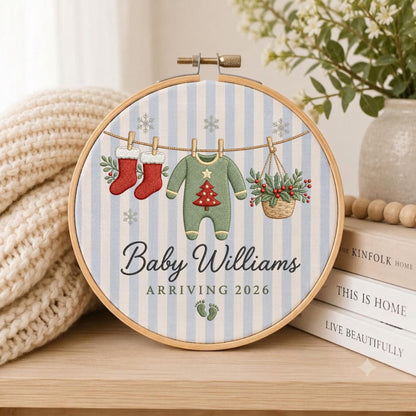 May include: A round wooden embroidery hoop featuring a light blue and white striped fabric background. The embroidery depicts two red stockings, a green baby onesie with a red Christmas tree, and a basket of holly hanging on a line with snowflakes.
